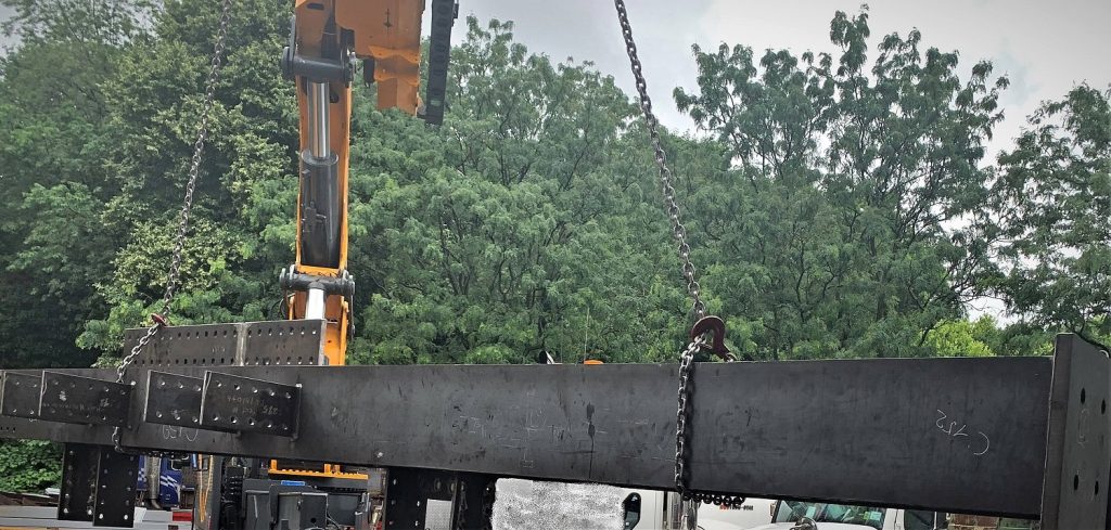 A steel girder with bolted connection plates hangs from chains on a crane hook above a truck, with trees behind.