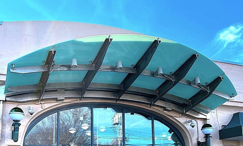 A curved green glass canopy is carried on a row of tapered steel outrigger arms bolted to a building face above a tall arched storefront window.