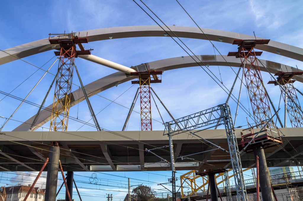 Two pale steel arch ribs of a bridge under construction rise on red and yellow temporary shoring towers above a steel deck girder and railway catenary wires, against a blue sky.