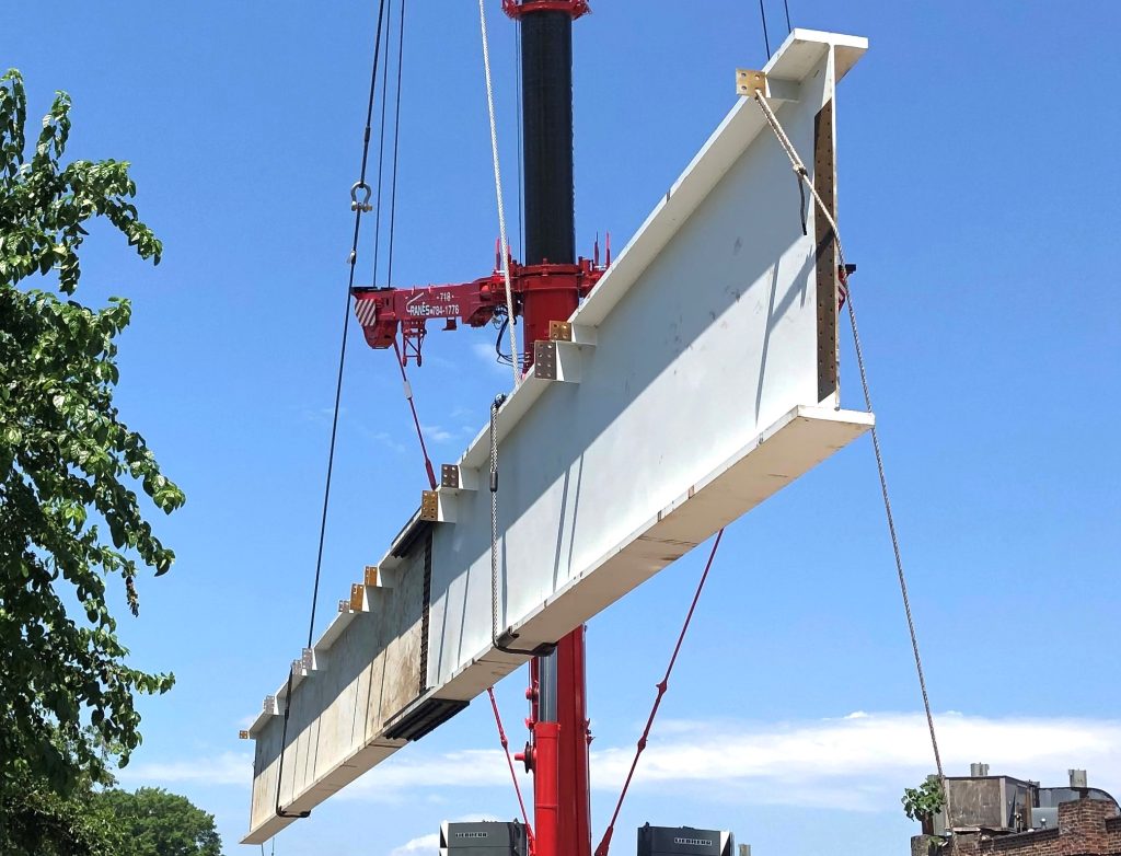 A long white painted steel plate girder with welded stiffeners hangs from crane slings against a blue sky, with the red crane boom behind it.
