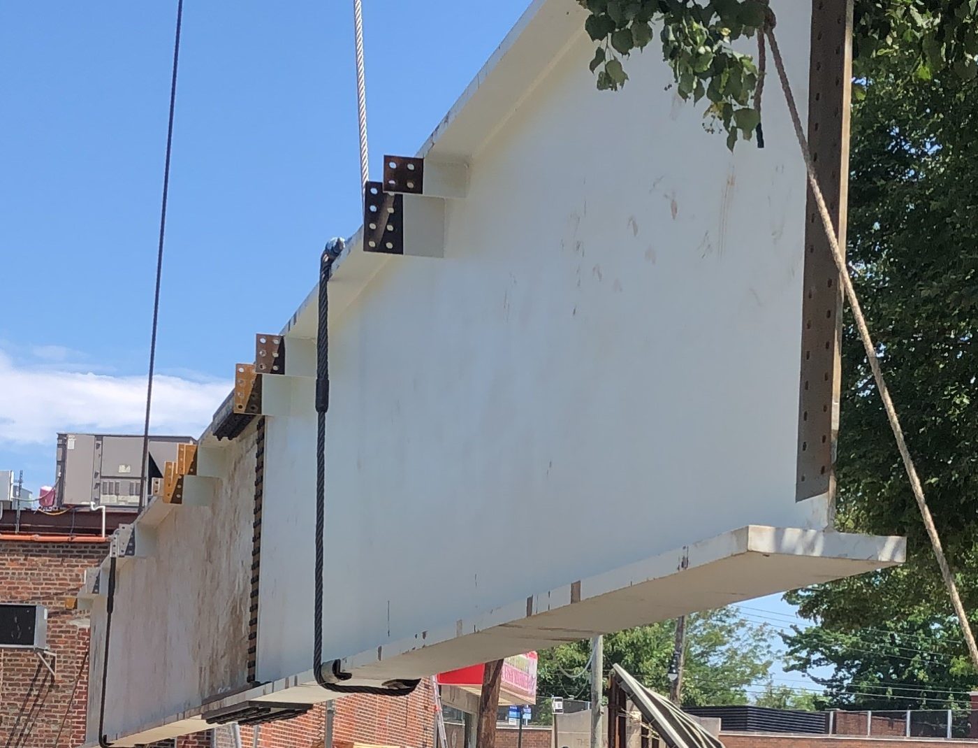 A white-primed steel plate girder with bolted connection plates hangs from wire rope slings above a brick street corner, trees behind.