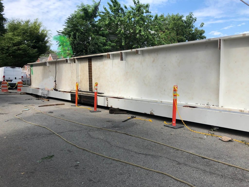 A long white-painted steel plate girder with a bolted field splice, blocked up on timber cribbing in a street closed off with orange delineators and traffic drums.