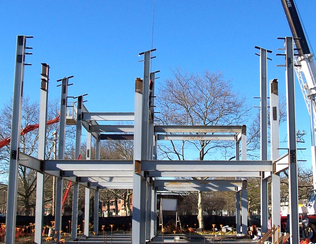 Painted steel columns and beams standing on a concrete slab with anchor bolts in the foreground and a crane at the right.