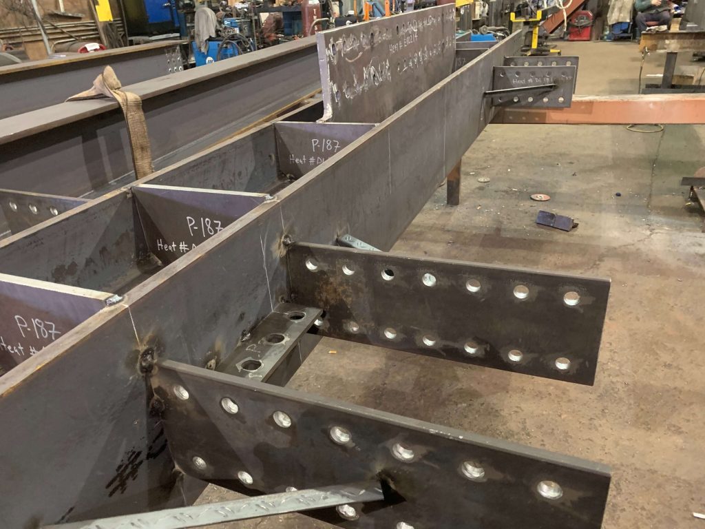 Raw steel girders with welded stiffeners and bolted splice plates on a shop floor, piece marks and heat numbers written in chalk.