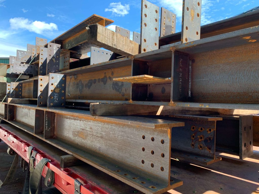 Fabricated steel beams with bolt-hole connection plates stacked on a red flatbed trailer outdoors against a blue sky with scattered clouds.