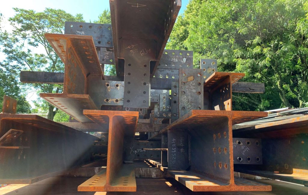 Low-angle view along a trailer loaded with fabricated steel beams and gusset plates, their bolt holes and stiffeners framed against trees and a bright sky.